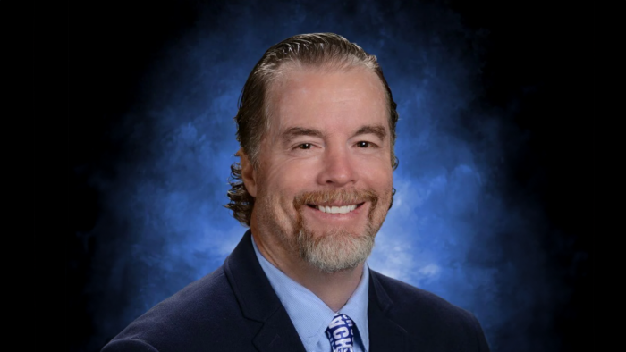 A professional headshot of A&M-Commerce alum Shae Creel in a dark suit and blue tie against a blue and black backdrop.