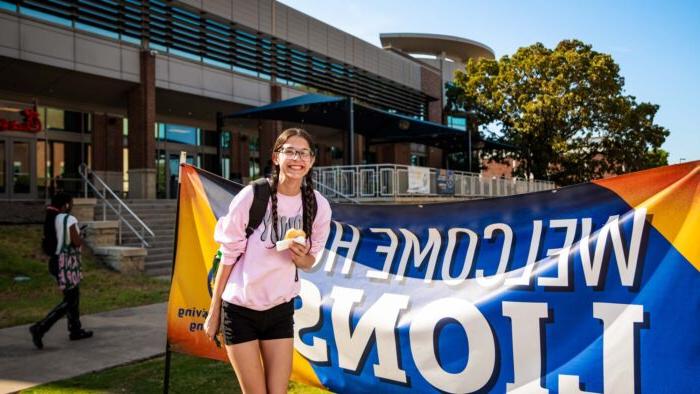 Student poses in front of a "Welcome Home Lions" banner, wearing shorts and a t-shirt and holding a donut.