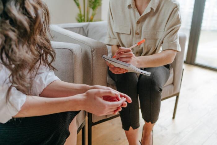 Two people sit sin chairs, representing a counseling session. Their faces are not in the frame. The photo shows the counselor writing on her notepad in the background and the patient with her hands clasped together on her lap in the foreground.